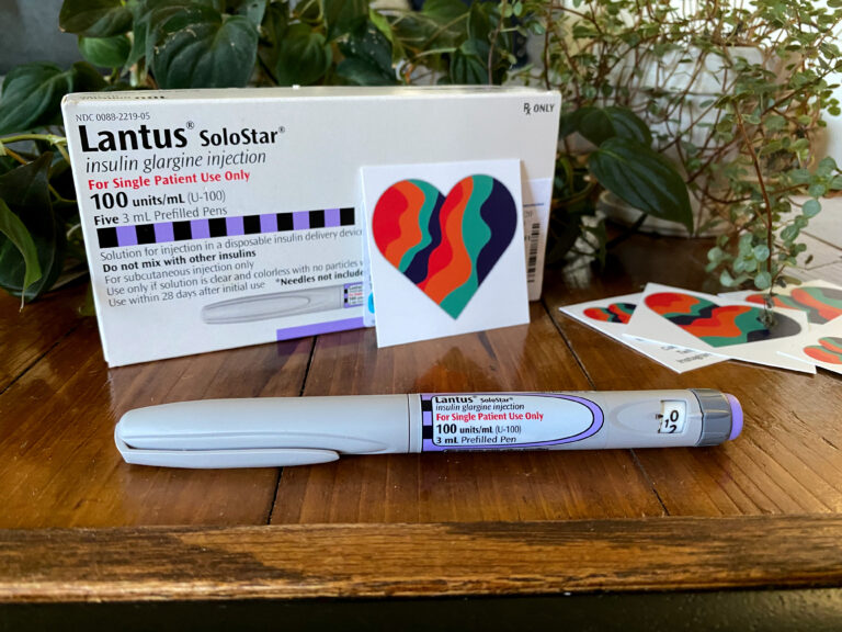Lantus pen on a wood table. Behind it is a box of lantus pens and MAD business cards with our red orange teal and dark blue heart logo